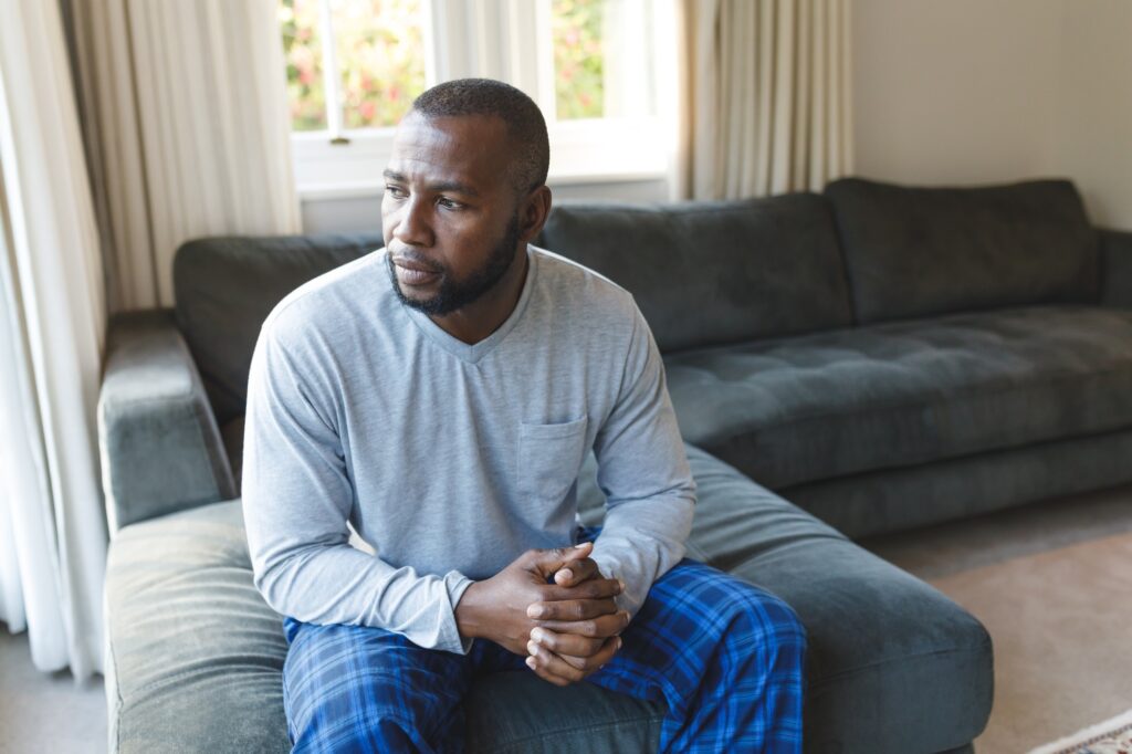 Worried, sad african american man thinking, sitting on couch looking out of window in living room