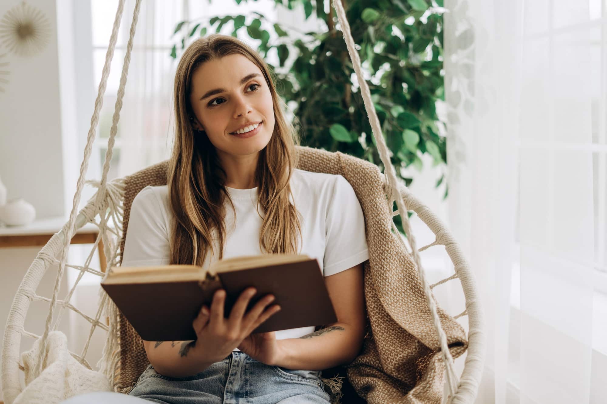 Young woman relaxing on hanging chair reading a book and looking away