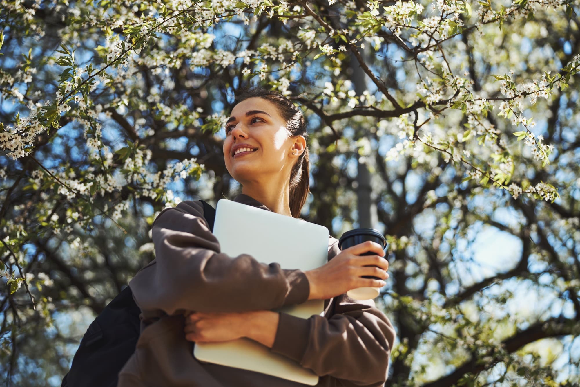 Hopeful lady bringing laptop and drinking coffee during walk outdoors